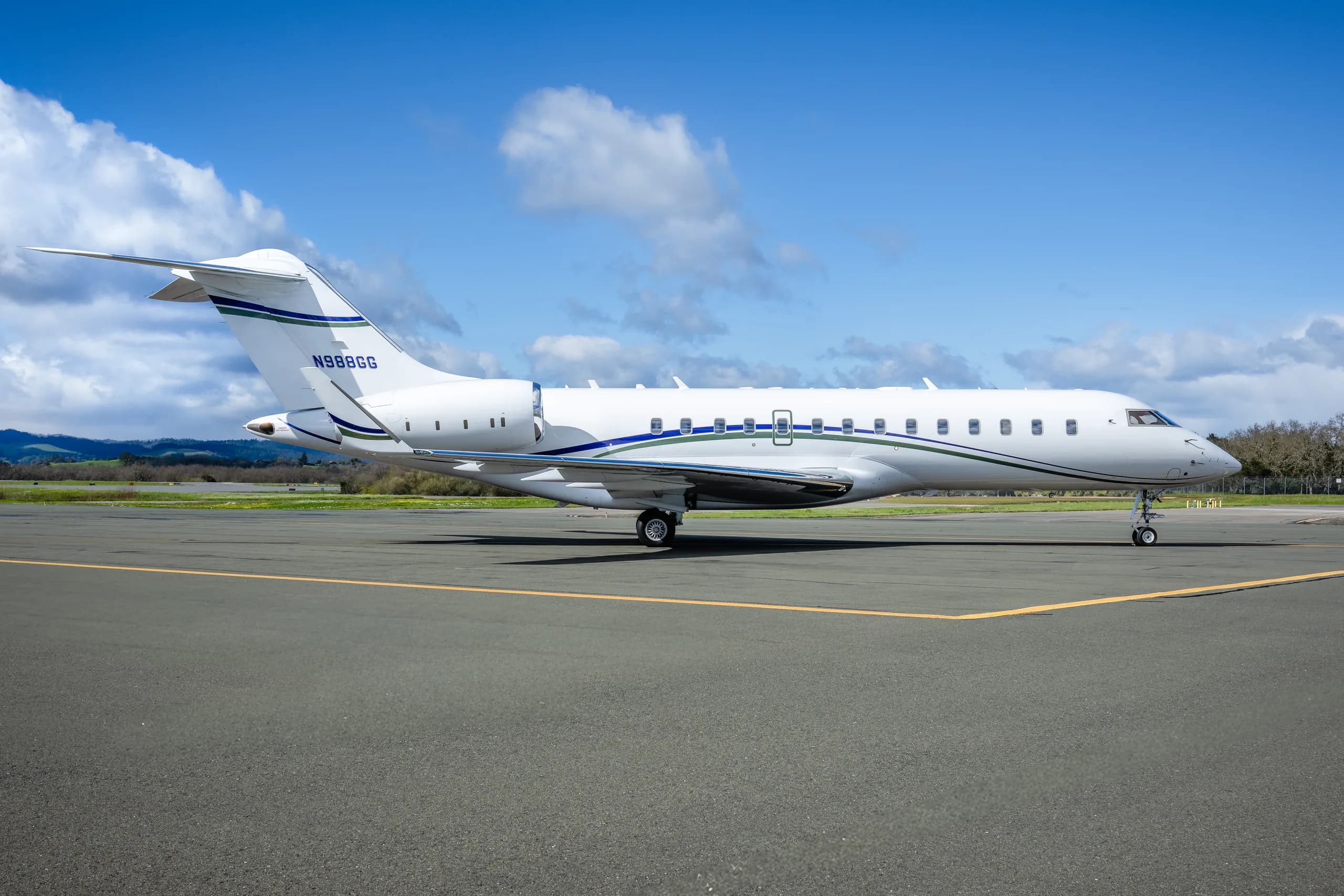 A white Bombardier Global 6000 business jet with blue and green stripes, parked on an airport tarmac with green fields and trees in the background.