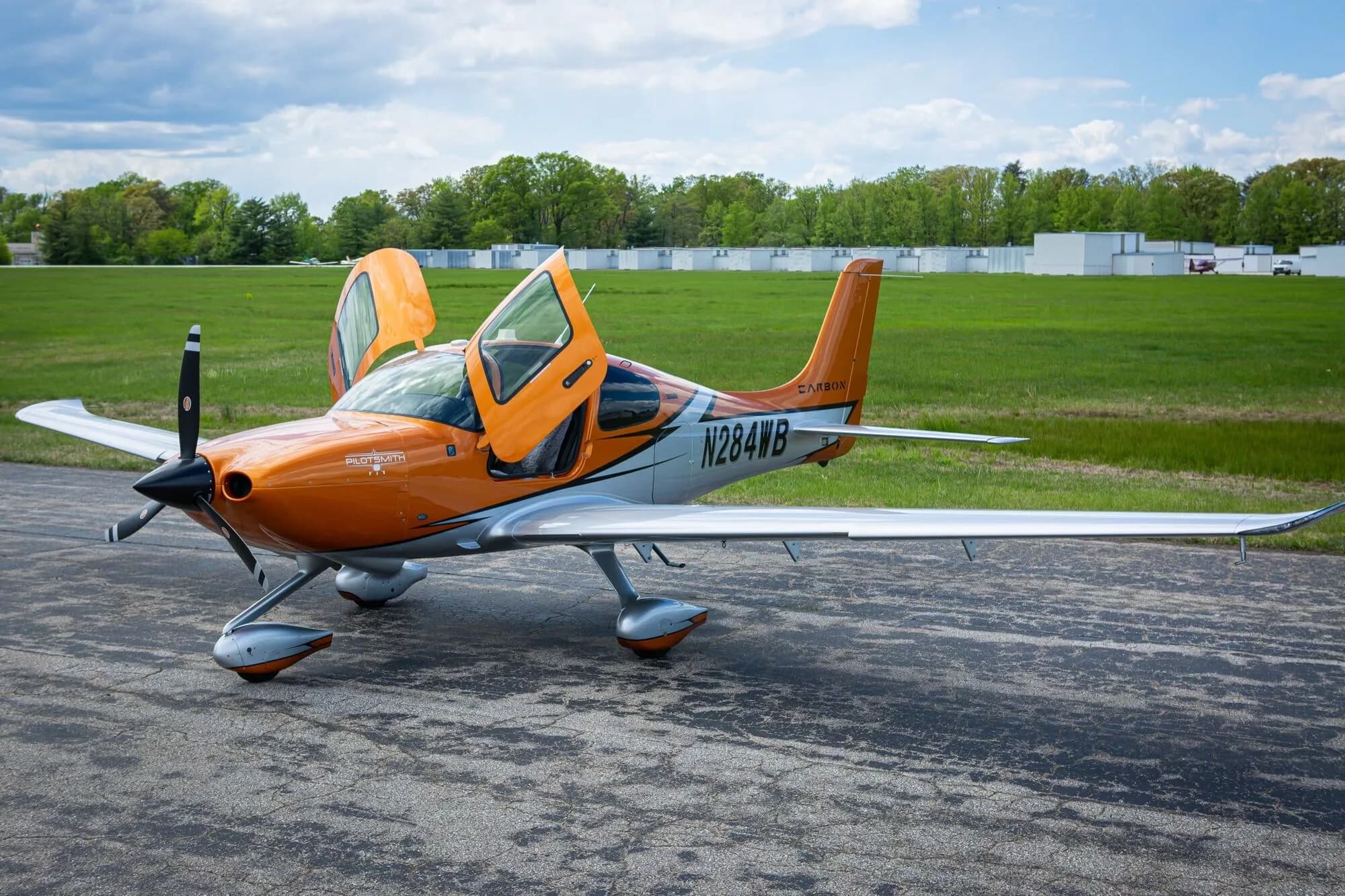 An angled front view of an orange and silver Cirrus SR20 aircraft, N284WB, with its gull-wing doors open on an airfield.