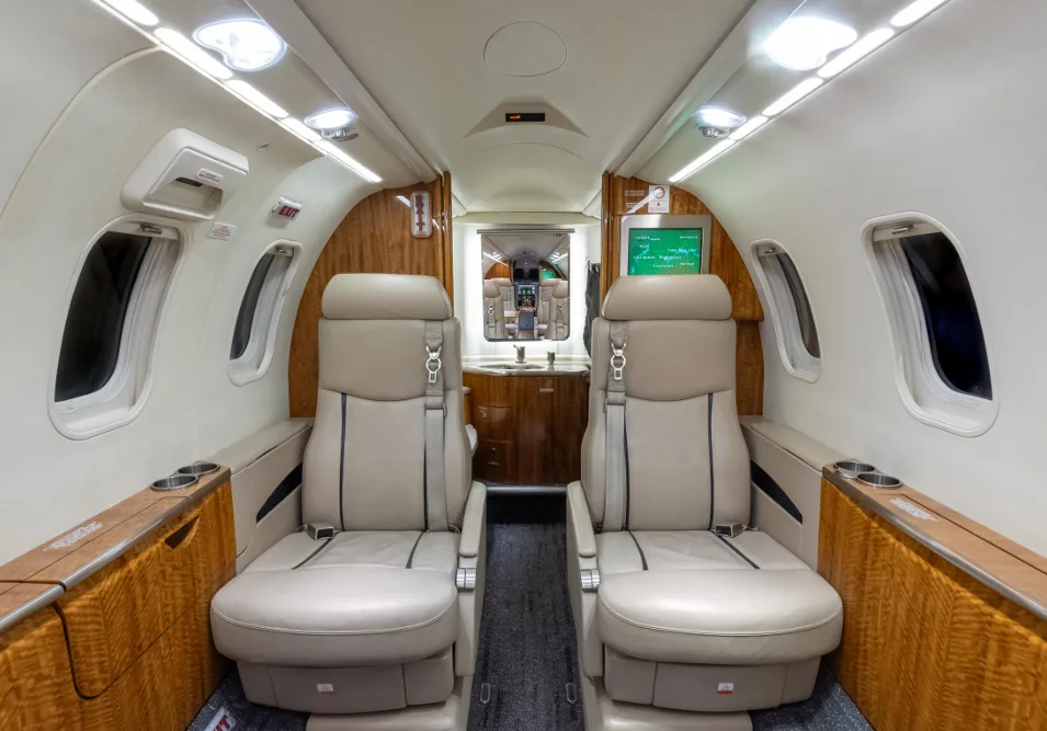 The rear section of a business jet cabin, showing beige seats, wood paneling, and a lavatory entrance.