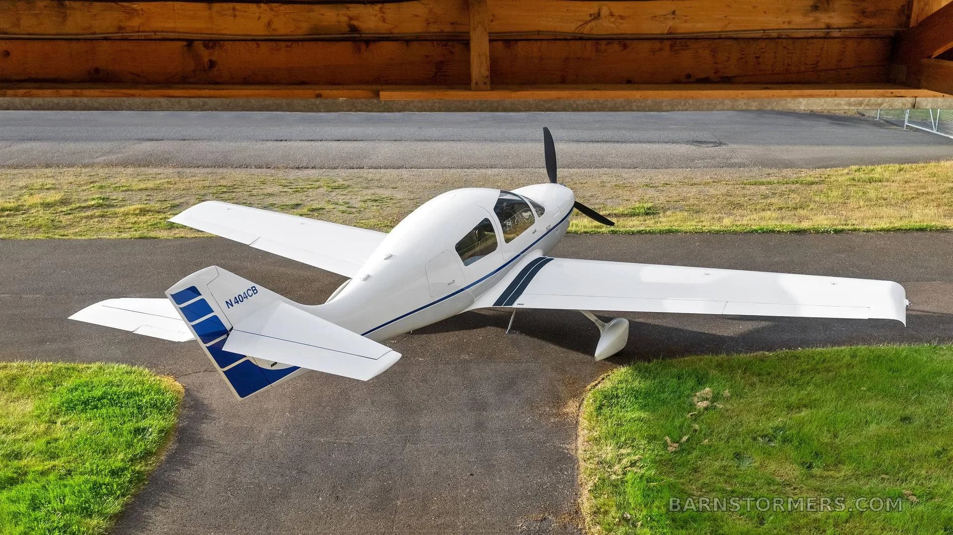 An overhead rear view of a white single-engine aircraft with blue stripes, registration N404CB, parked on an asphalt surface near a wooden structure.