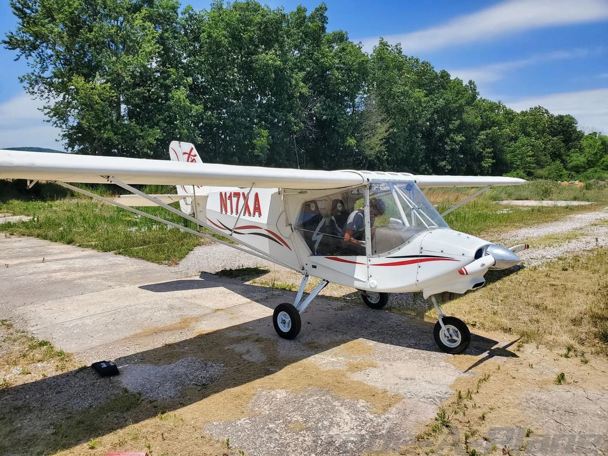 A white X-Air aircraft with red and black stripes, registration N17XA, is parked on a concrete pad with trees in the background.