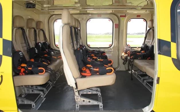 Interior view of a helicopter cabin showing multiple rows of beige seats with harnesses, looking towards the rear.