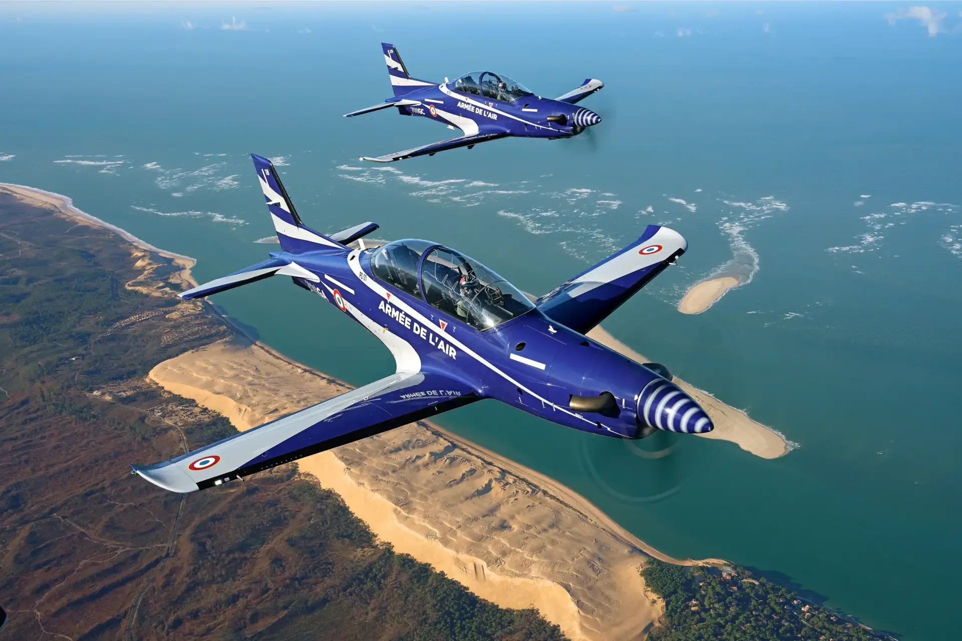 Two blue and white Pilatus PC-21 aircraft flying in formation over a coastal landscape.