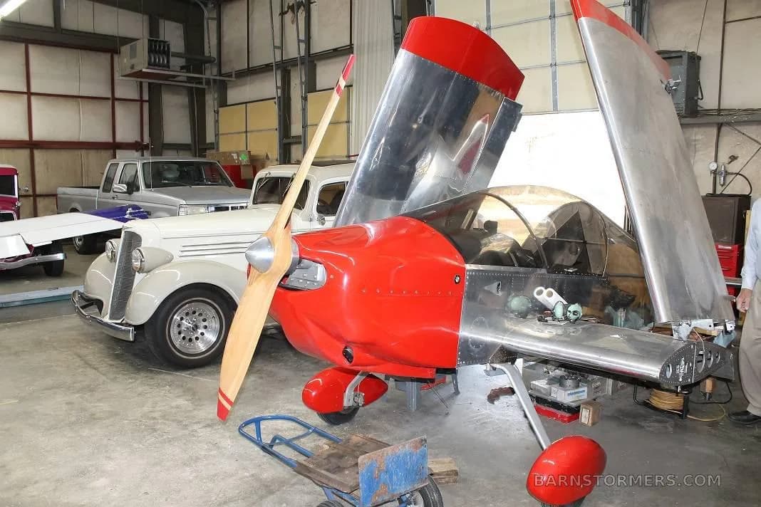 A silver and red experimental aircraft with open wings is parked next to classic cars in a workshop.