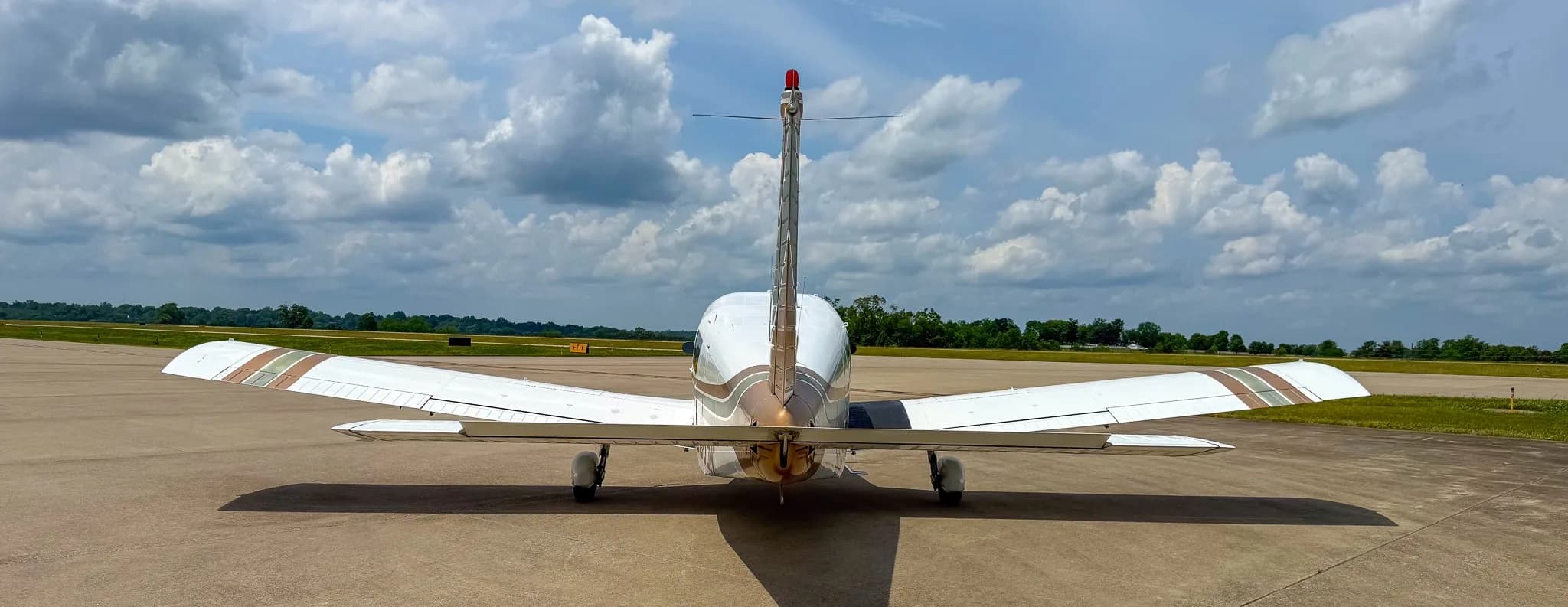 A rear view of a white Piper Cherokee Six aircraft with brown and green stripes parked on an airfield.