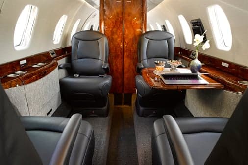 A private jet cabin interior showing black leather seats, a wooden table with a laptop and drinks, and elegant wood paneling.