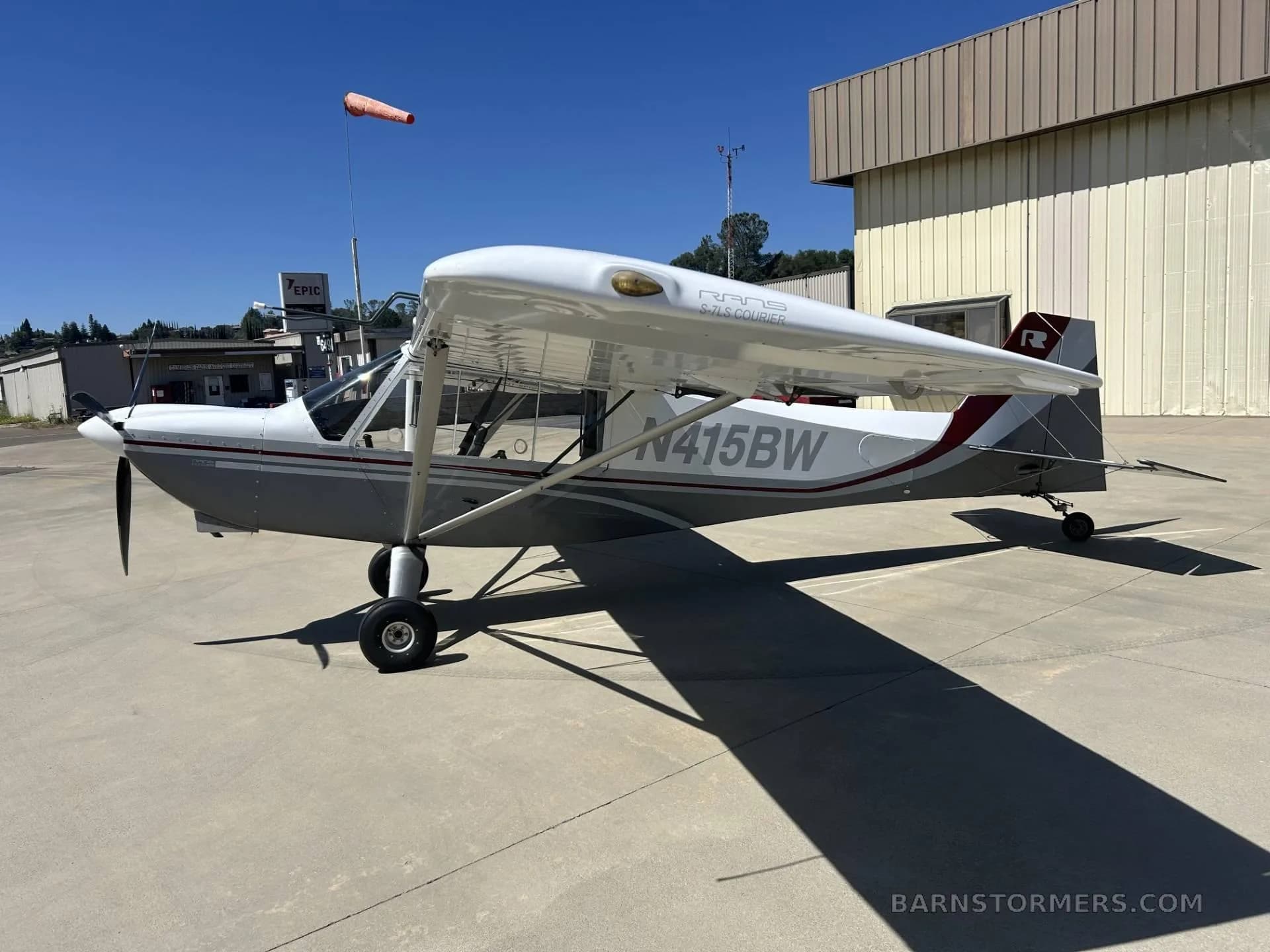 A side view of a white and grey Rans S-21 Outbound aircraft, N415BW, parked on a sunny tarmac with a windsock and buildings in the background.