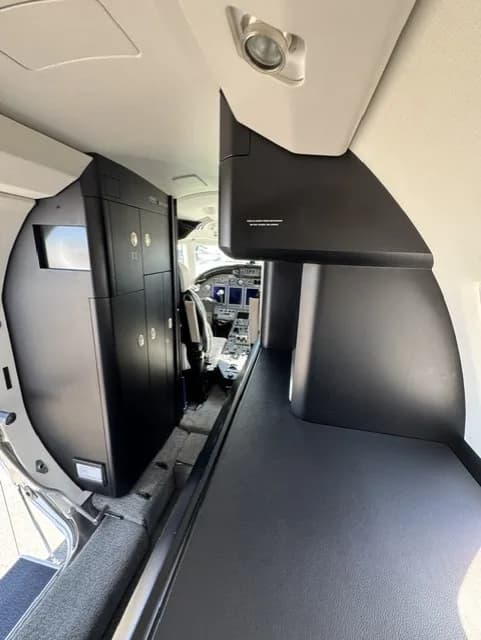 The interior of a business jet, showing dark cabinetry along the side and a glimpse of the cockpit.