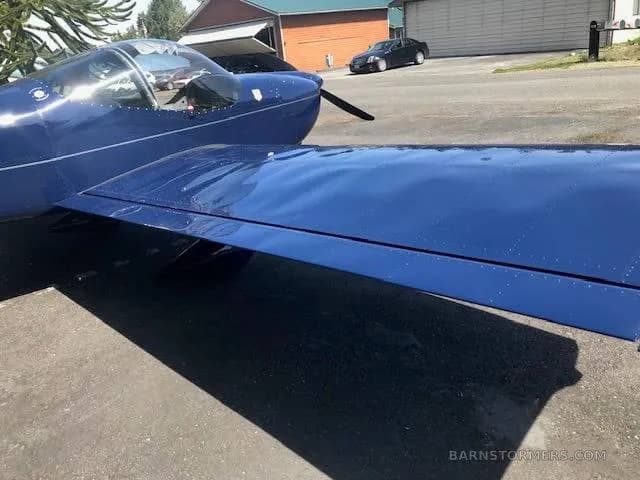 A close-up view of the dark blue wing and cockpit of an aircraft, with a car in the background.