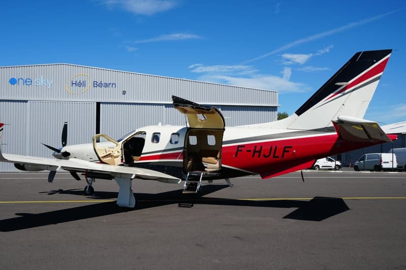 A TBM 930 aircraft with its door open is parked in front of a hangar.