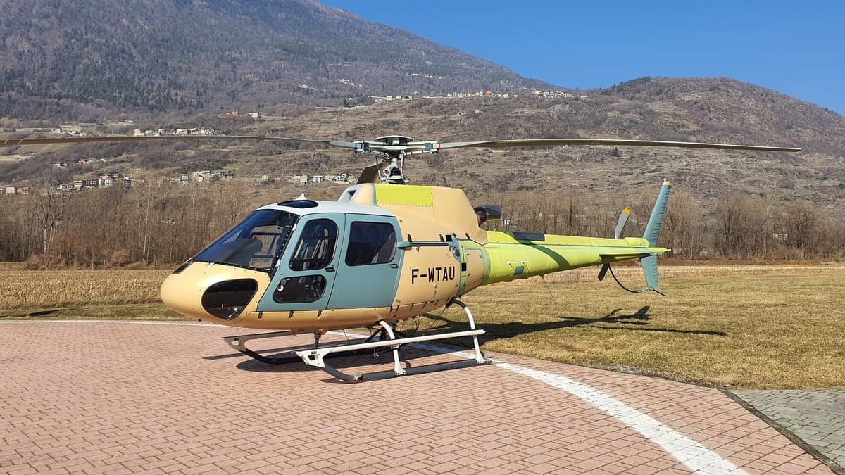 A Eurocopter AS350 Ecureuil helicopter is parked on a helipad with mountains in the background.