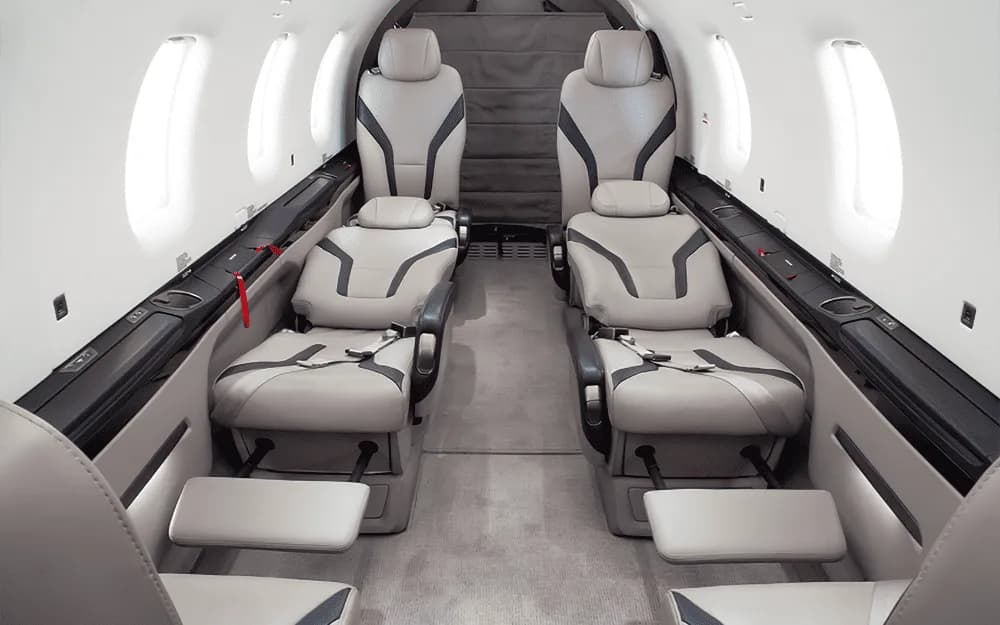 A wide view of the Pilatus PC-12 aircraft cabin, showing multiple rows of light grey seats with footrests.