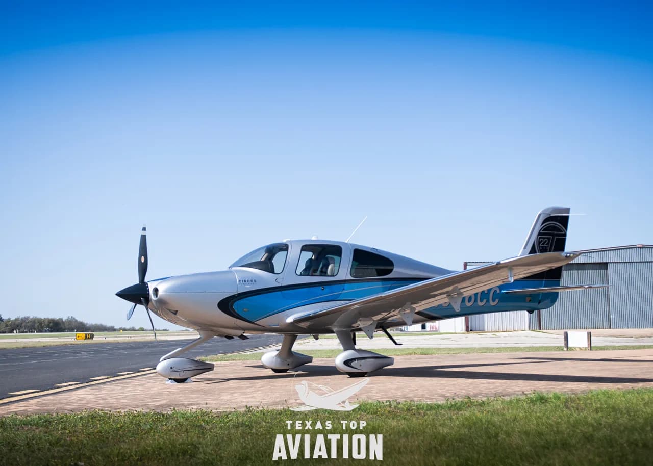 A Cirrus SR22 aircraft with blue and silver paint scheme parked on an airfield under a clear blue sky.