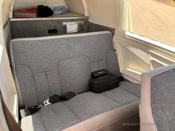 The rear cabin of a small aircraft features a grey fabric bench seat with seatbelts and a small storage area.