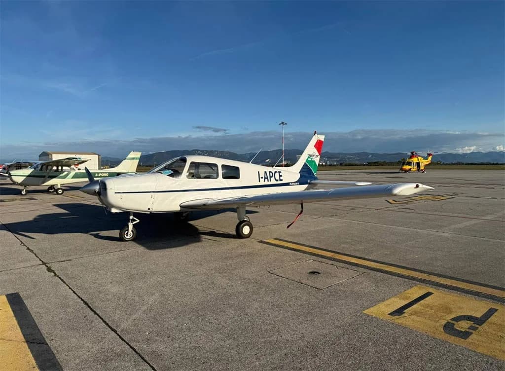 A white single-engine propeller aircraft with "I-APCE" registration parked on an airfield with other aircraft in the background.