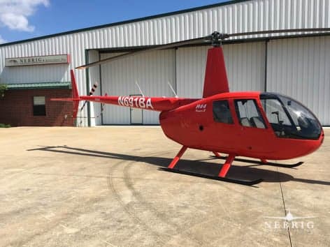 A red Robinson R44 Raven II helicopter with registration N697BA parked on a concrete pad in front of a white hangar.