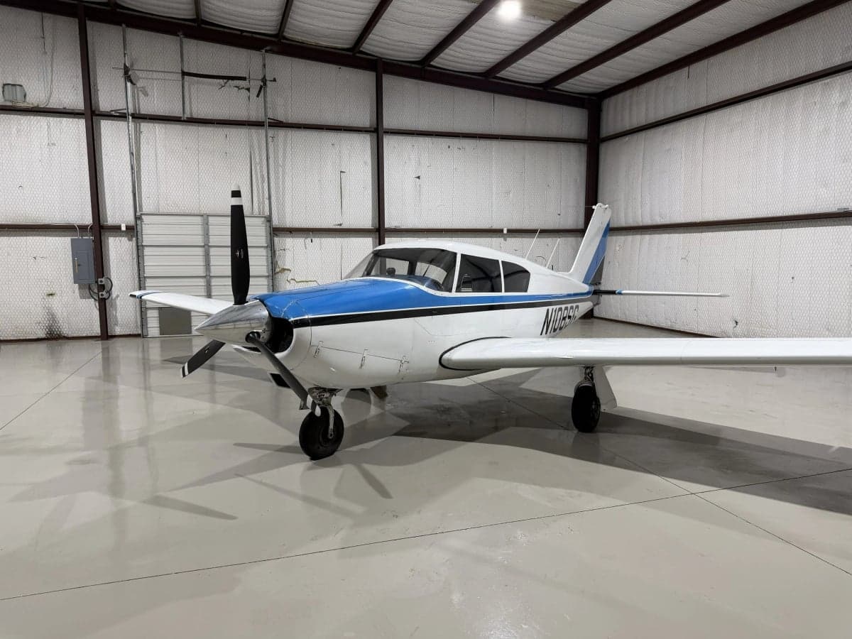 A white and blue Piper PA-28-140 Cherokee is parked in a hangar.