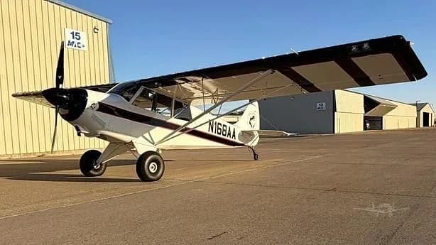A white and black high-wing single-engine aircraft, N168AA, is parked on an airfield with hangars in the background.