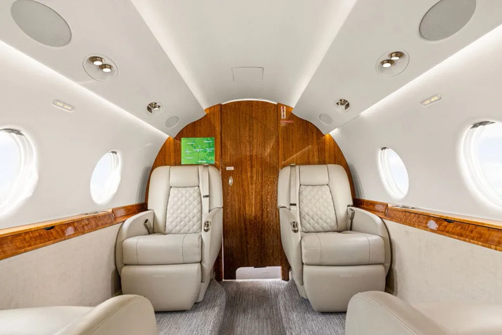 The luxurious interior of a private jet cabin features two beige quilted seats facing a wooden bulkhead.