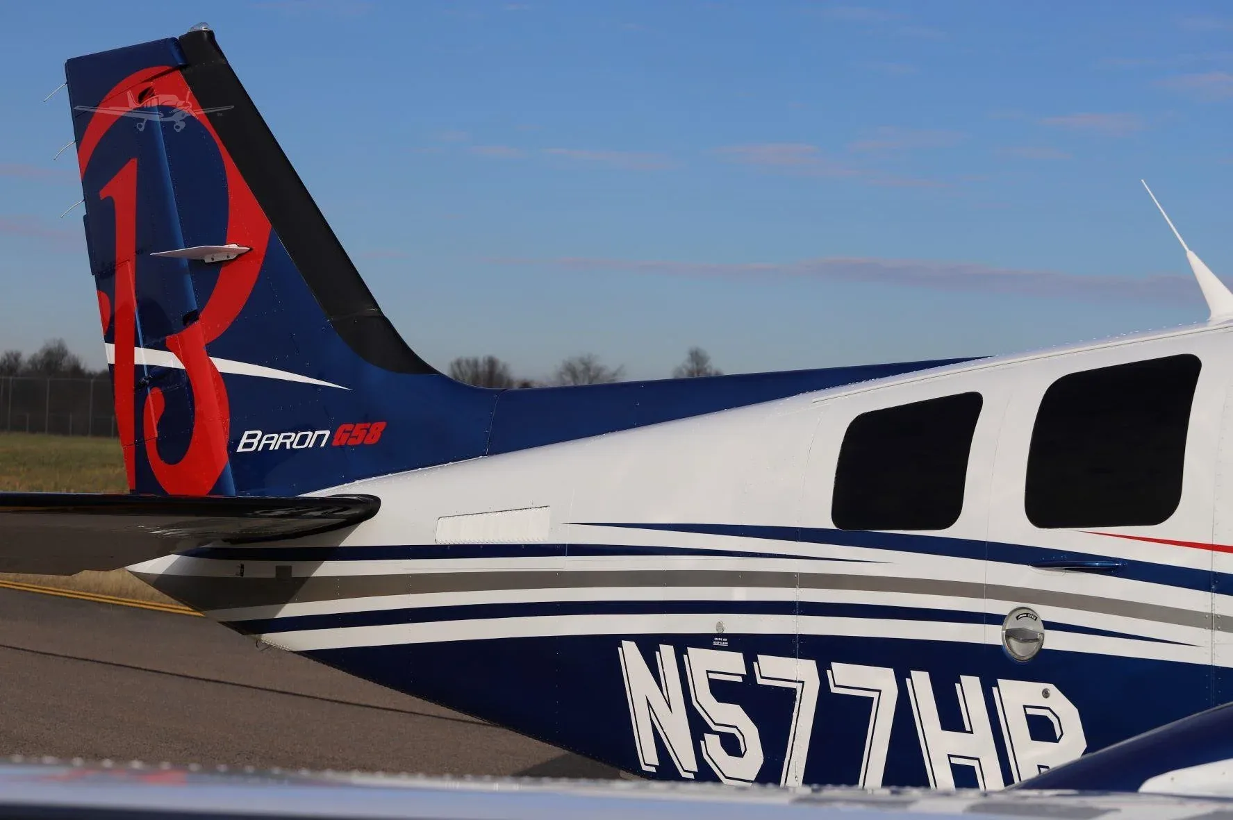 A close-up of the tail and rear fuselage of a white and blue Beechcraft Baron G58 (N577HR) with a large red 'B' logo on the tail.