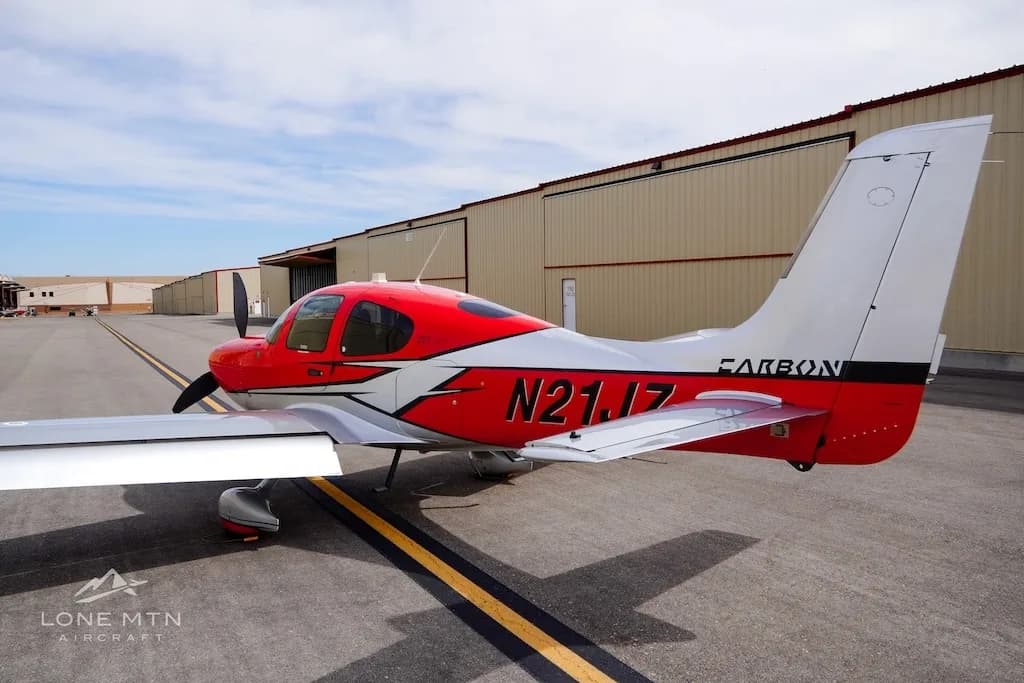 A rear-side view of a red and silver Cirrus SR22 aircraft parked on an asphalt ramp with hangars in the background.