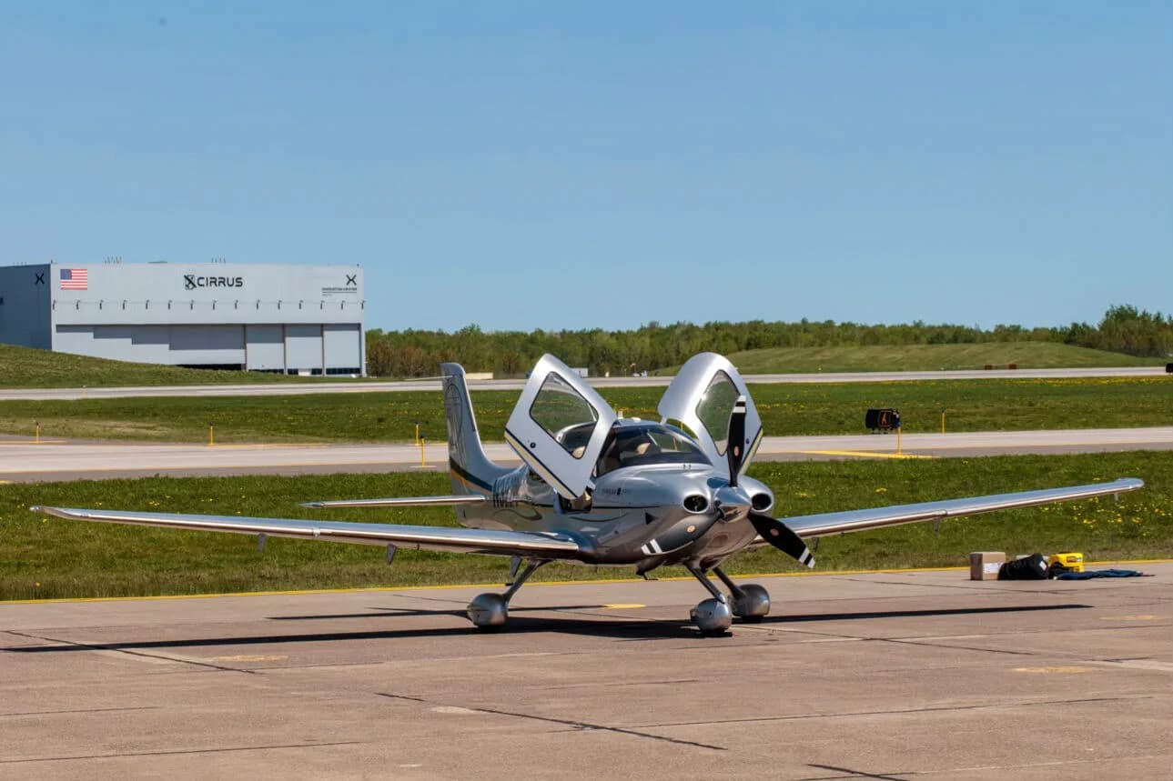 A silver Cirrus SR22T aircraft with its gull-wing doors open, parked on an airfield with a Cirrus hangar in the background.