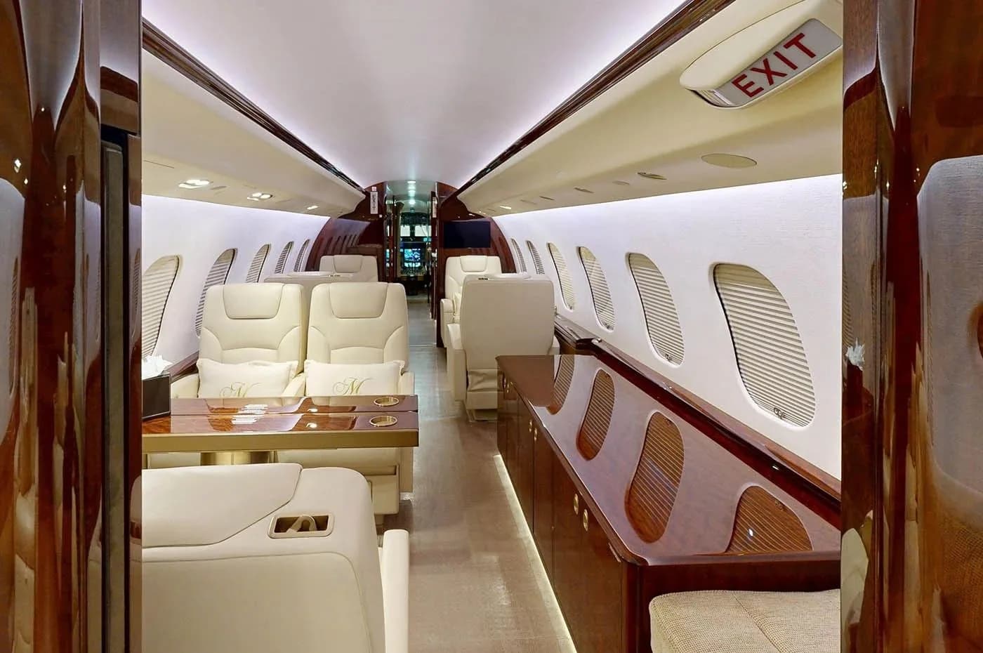 A view down the private jet cabin, showing cream-colored seating, a polished wooden table, and dark wood paneling along the walls.