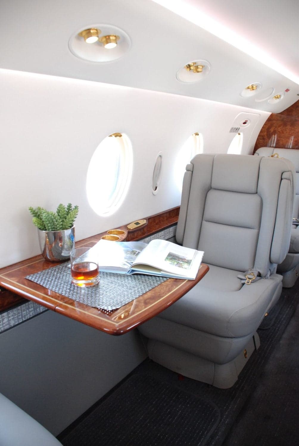 A comfortable grey leather aircraft seat with a fold-out table holding a drink and a magazine next to a window.