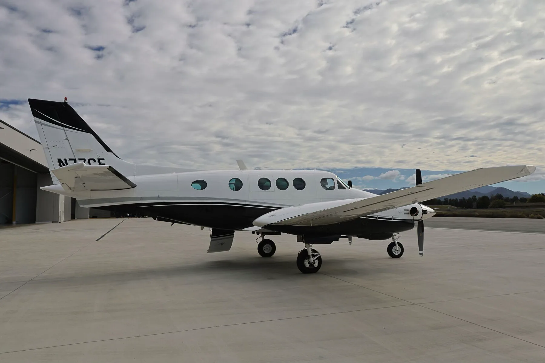 A left side view of the N77CE twin-engine turboprop aircraft parked on an airfield under a cloudy sky.