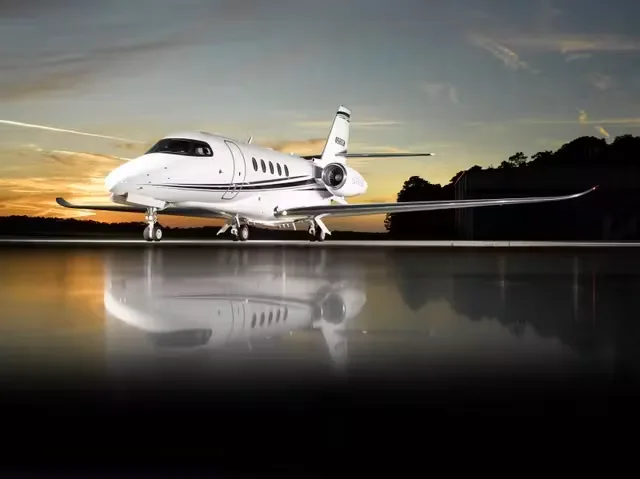 A white business jet with blue and grey stripes parked on a wet tarmac at sunset, reflecting in the water.