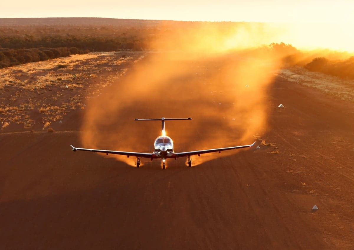 A Pilatus PC-12 aircraft takes off from a dusty runway at sunset.