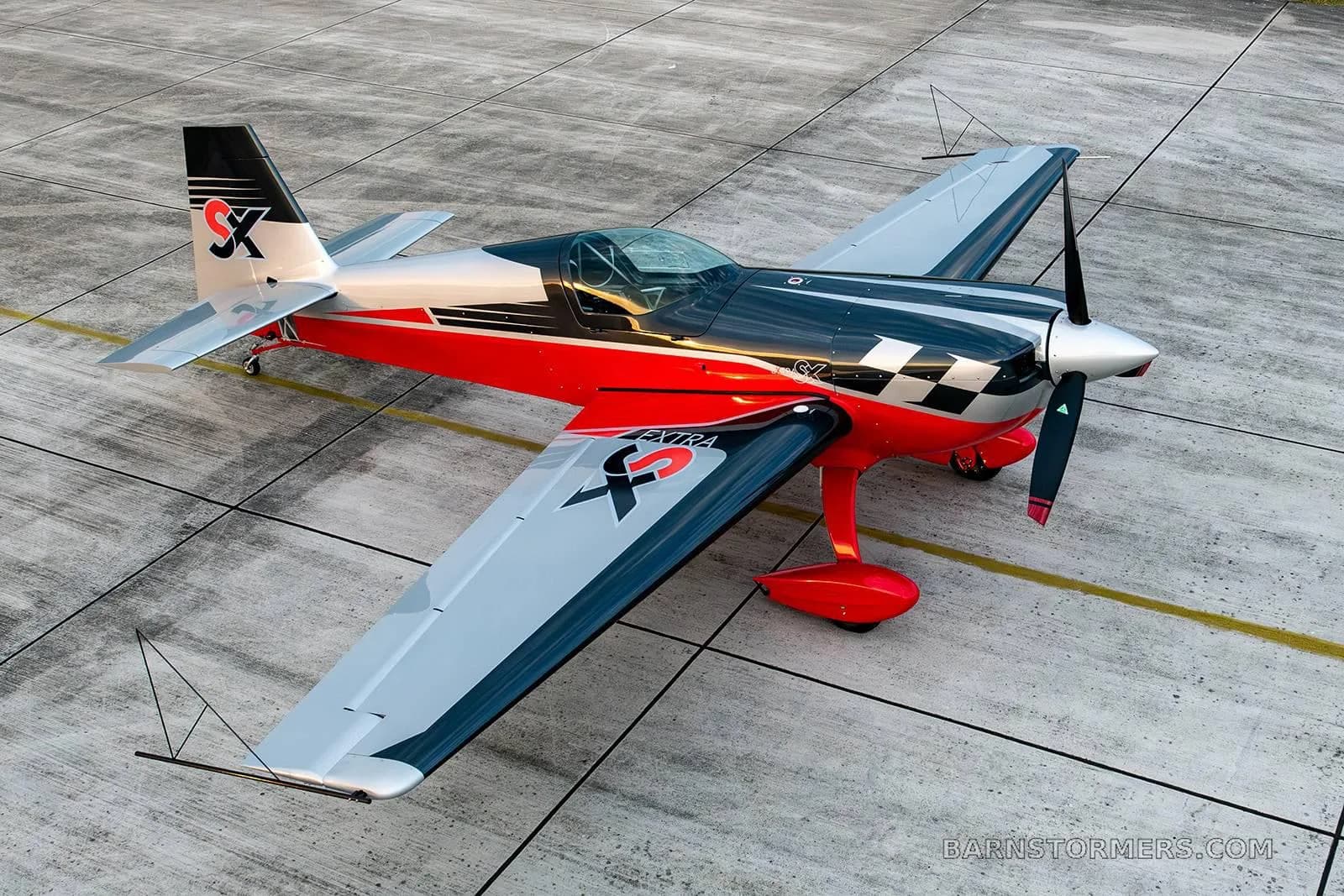 A high-angle view of a red, black, and silver Extra 330SC aerobatic aircraft parked on a concrete apron.