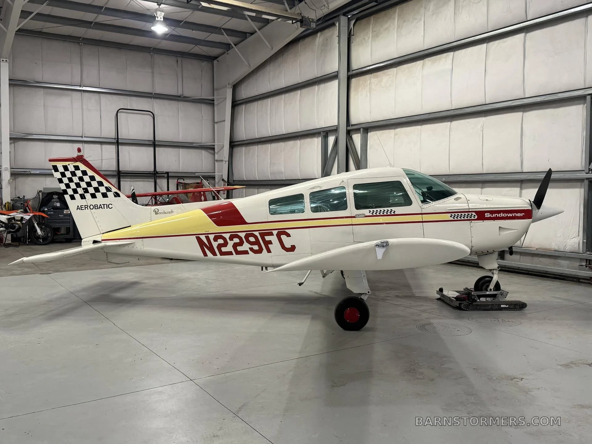 A white and red Beechcraft Sundowner N229FC with a checkered tail is parked in a hangar, viewed from the side.
