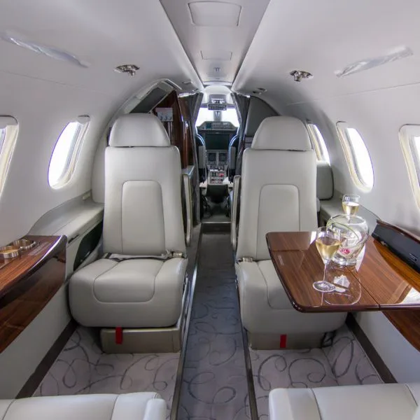 View of a private jet cabin with light grey seats, a wooden table with champagne, and a glimpse of the cockpit.