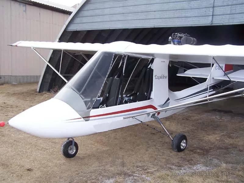 A white experimental aircraft with red and gray stripes, parked outside a hangar on a dirt ground.