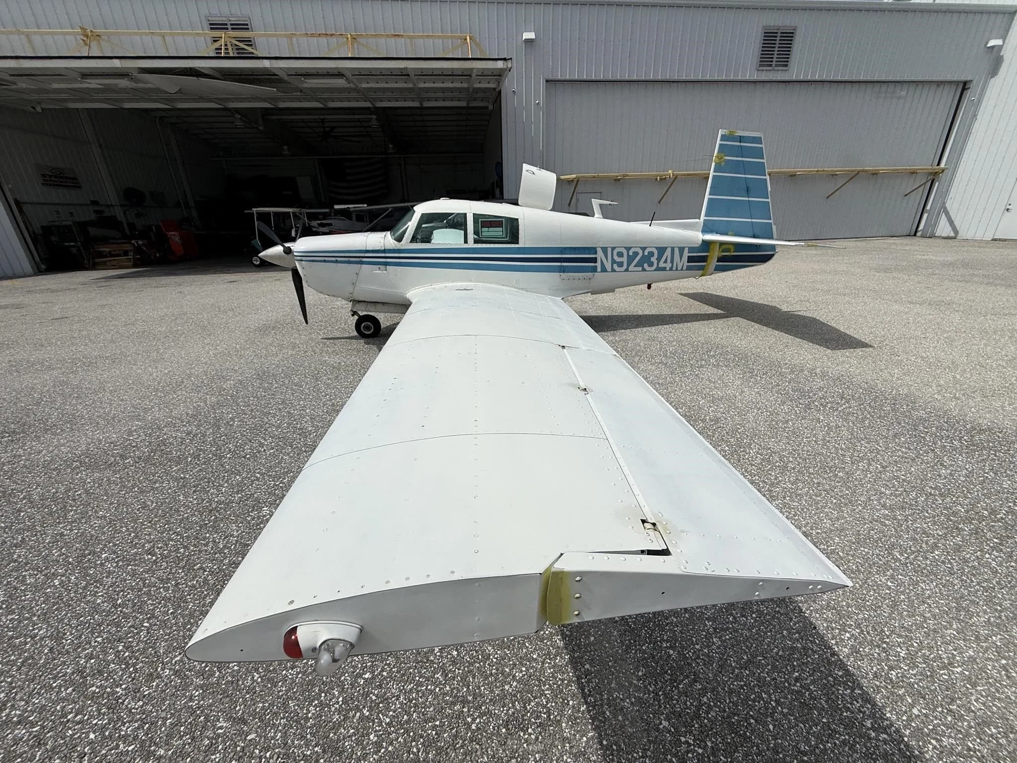 A white single-engine aircraft with blue stripes and registration N9234M, parked on an asphalt apron in front of an open hangar.