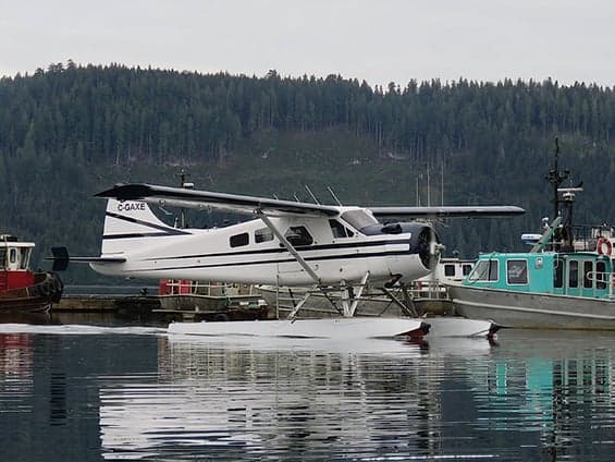 A white and blue floatplane is docked next to a boat on a lake.