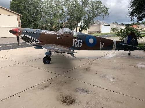 A P-40 Warhawk replica aircraft with shark mouth paint scheme is parked on a concrete pad.