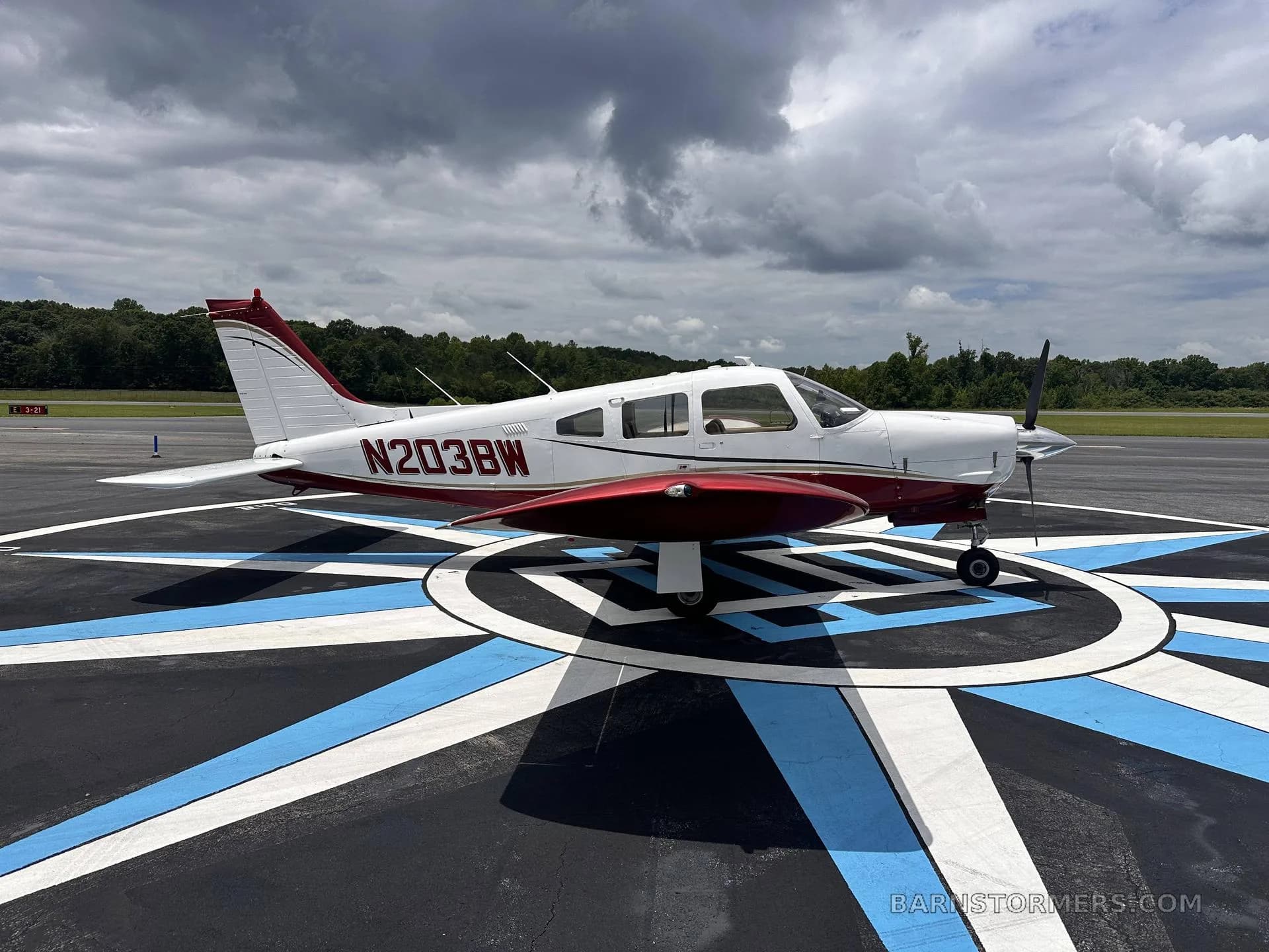 A white and maroon single-engine propeller aircraft (N203BW) is parked on a blue and white patterned tarmac under a cloudy sky.