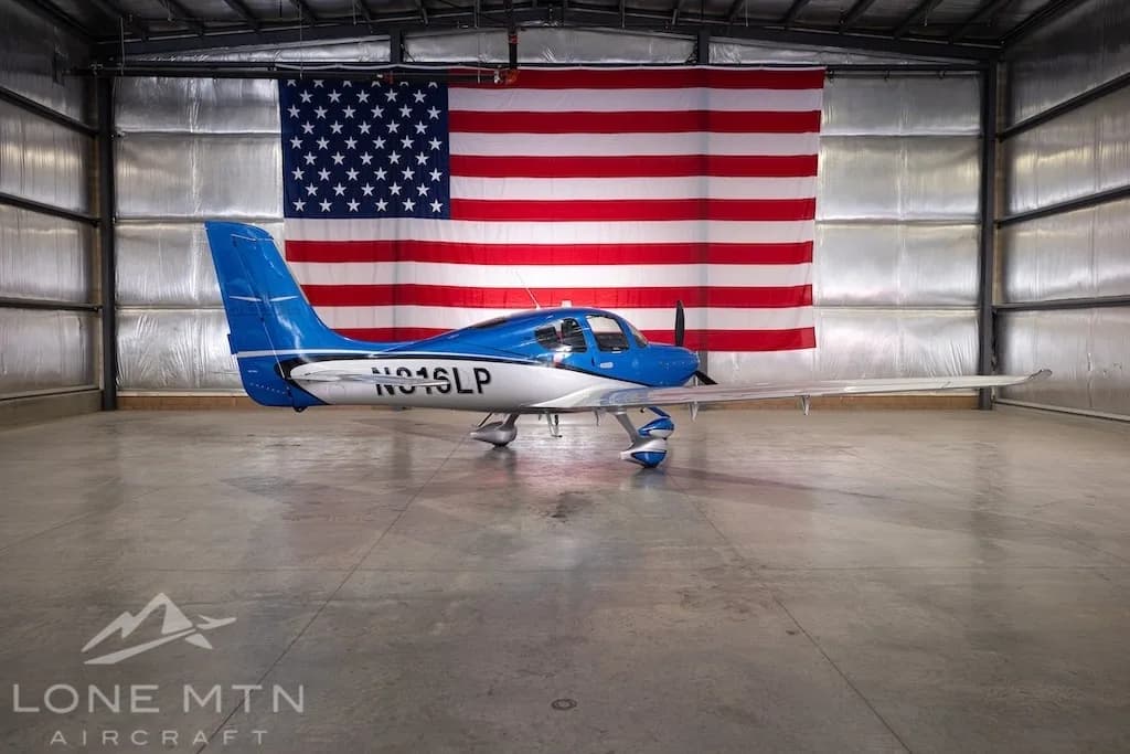 A blue and silver Cirrus SR22 aircraft, registration N816LP, parked in a hangar with an American flag in the background.