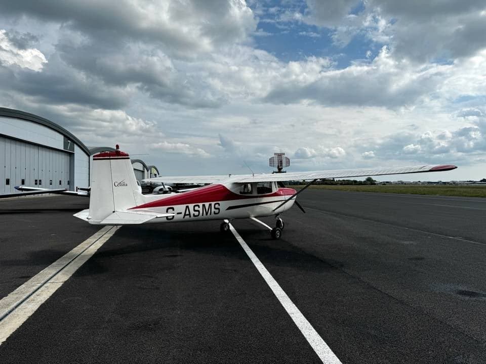 A white and red Cessna 150 G-ASMS parked on the tarmac, viewed from the rear-left, with an airport beacon in the background.