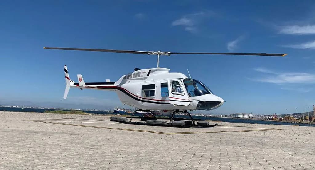 A white helicopter with black and red stripes on a helipad next to a body of water under a bright blue sky.