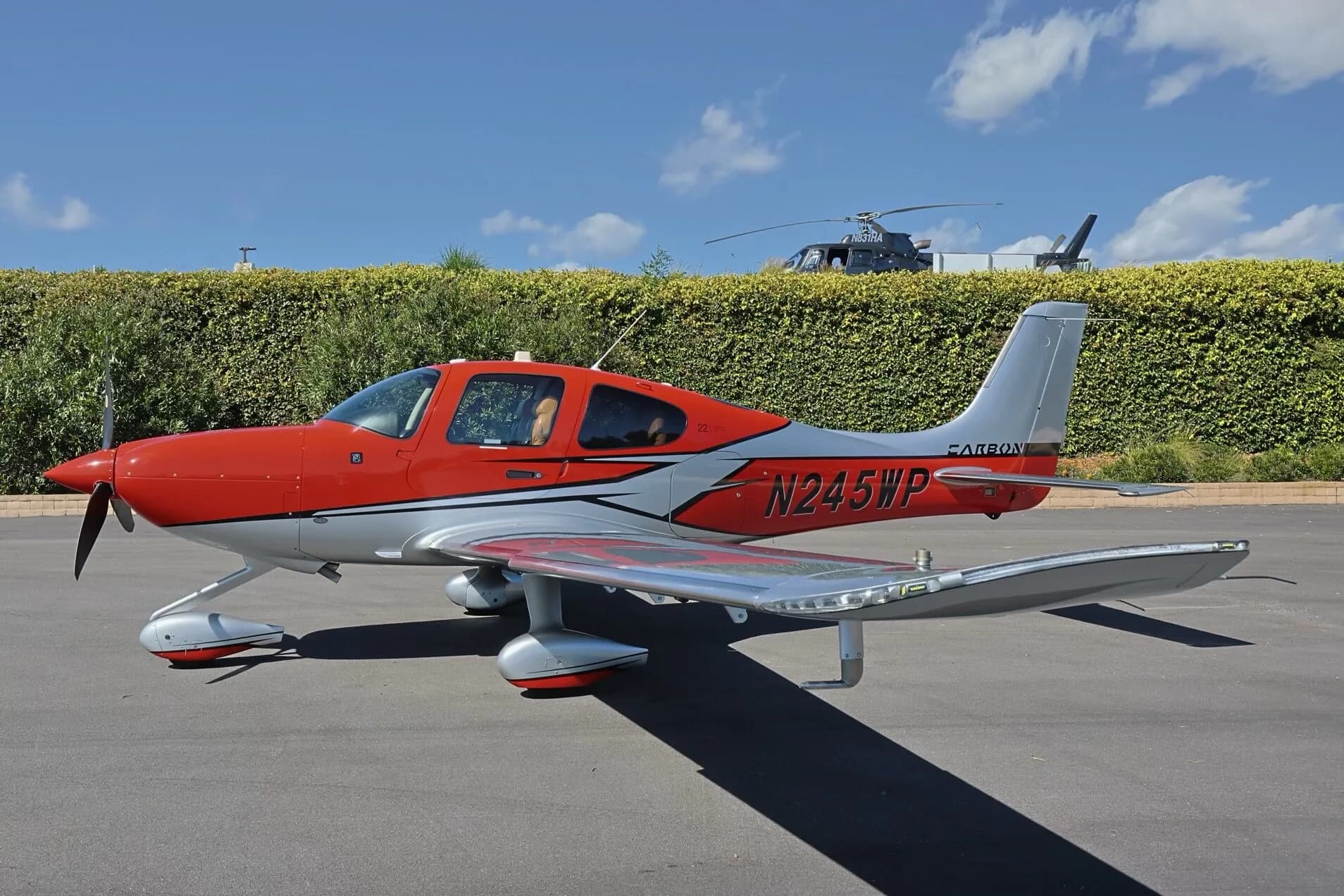 A red and silver Cirrus SR22 GTS aircraft, N245WP, parked on an tarmac with a helicopter partially visible.
