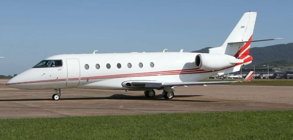 A white business jet with red stripes on the fuselage and tail, parked on a sunny tarmac with green grass in the foreground.