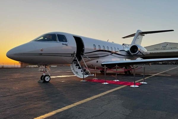 A white private jet with a red carpet leading to its open door is parked on an airfield at sunset.