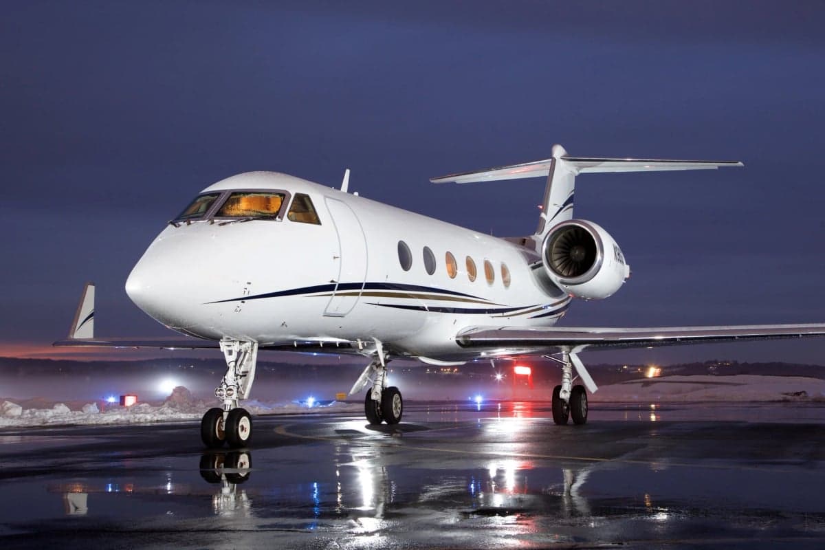 A white Gulfstream G450 business jet with gold and blue stripes parked on a wet tarmac at night, reflecting lights.