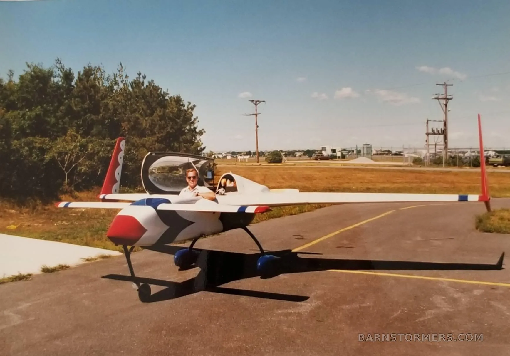 A white, red, and blue aircraft with twin tails and a pusher propeller, parked on an asphalt runway with a pilot in the open cockpit.