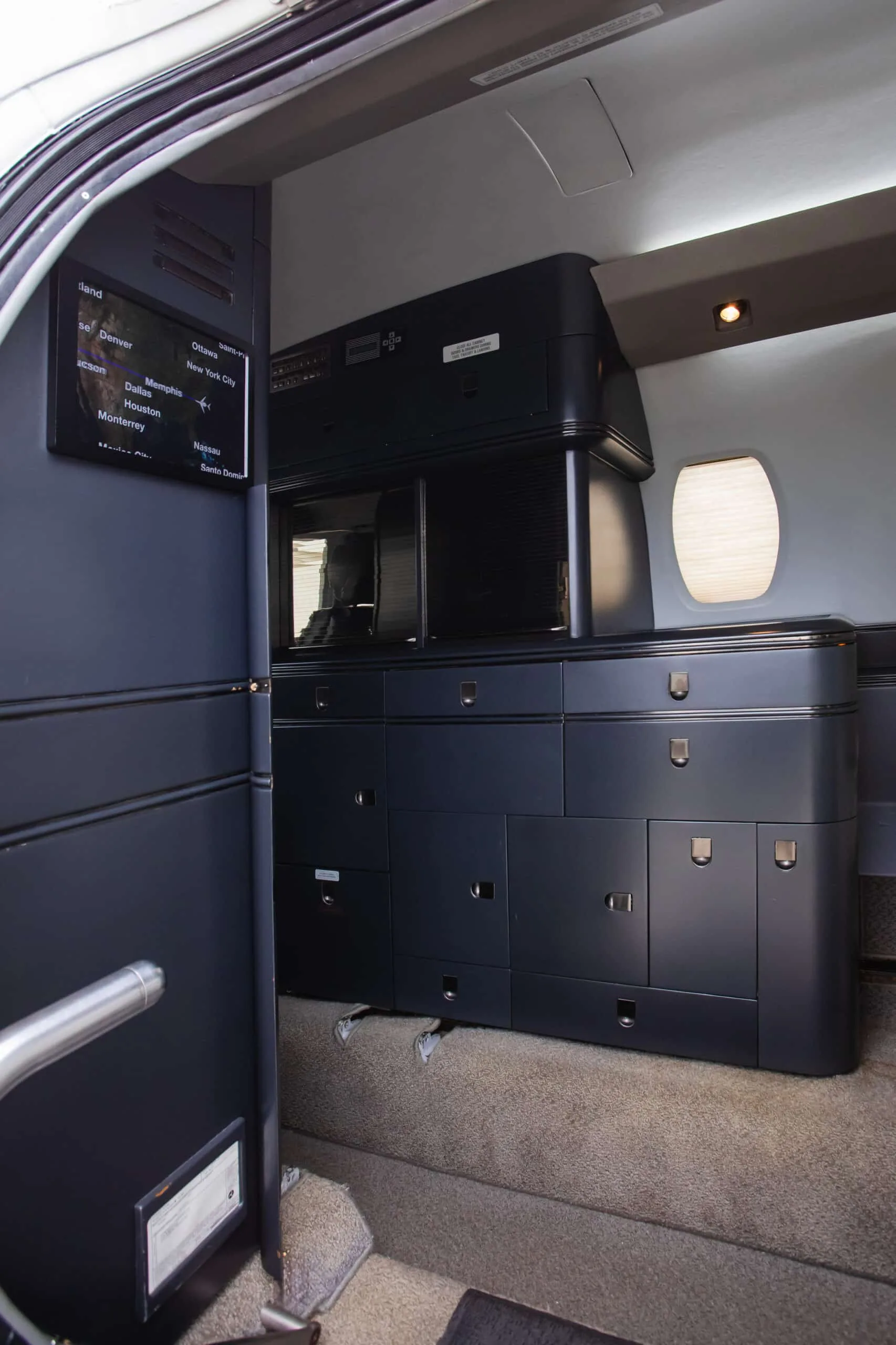 The galley or storage area of a private jet cabin with dark cabinetry and a screen displaying flight information.