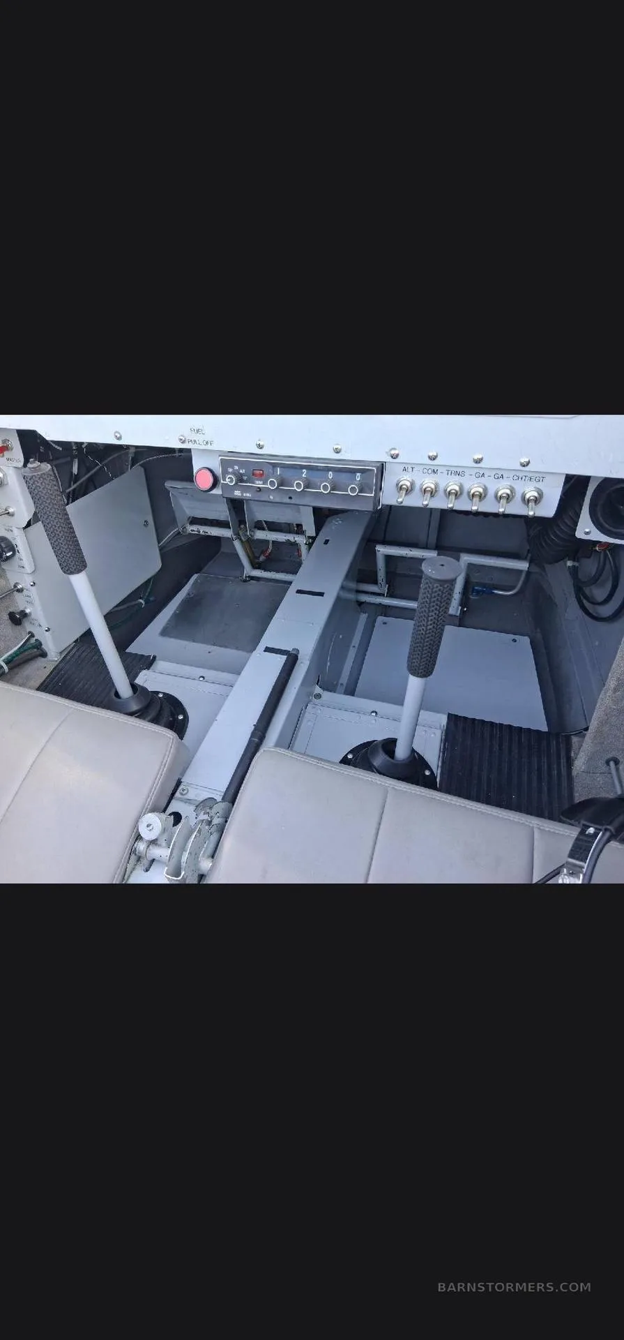 The interior cabin of a small aircraft, showing two light-colored seats and two control sticks on the floor.