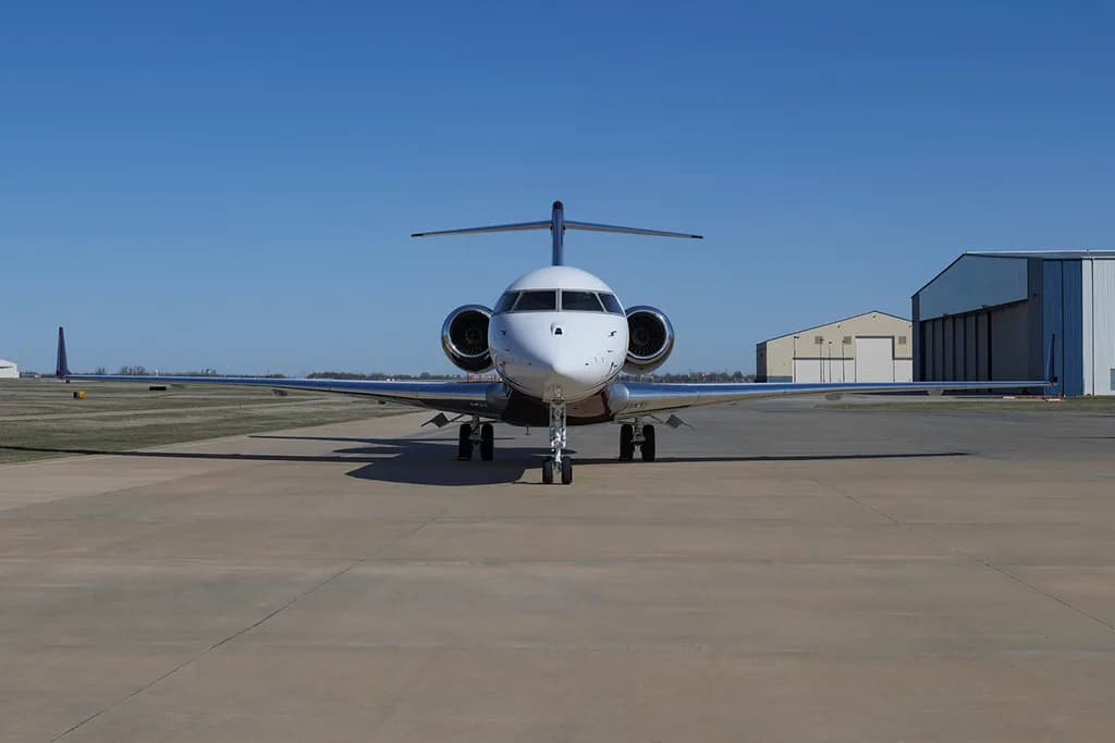 A Bombardier Global Express business jet with a white and maroon paint scheme, registration N919EM, viewed head-on on a tarmac with hangars in the background.